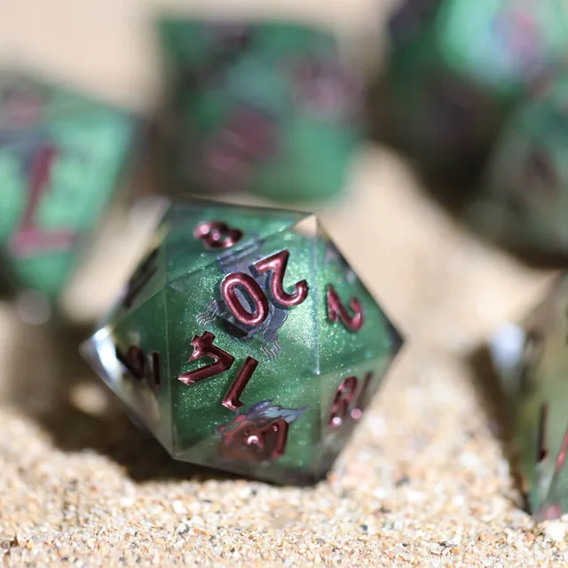 Green D20 on sand with out-of-focus dice behind it.