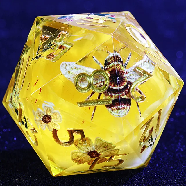 Close-up of a golden polyhedral die with a bee, flowers, and gold-coloured numbers.